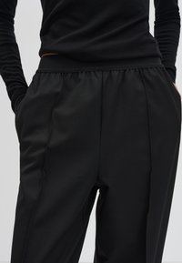 Pantalons de jogging noirs avec une finition lisse, taille élastique, poches latérales et design de jambe droite ajustée. La texture du tissu doux est visible.