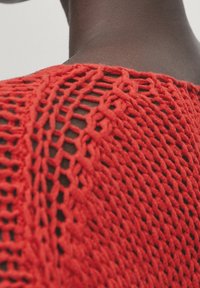 Haut rouge en maille avec un motif à mailles ouvertes, présentant une finition texturée le long du cou et de la zone des épaules.