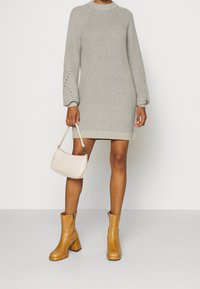 Abito a maglia grigio chiaro con texture a coste, maniche lunghe e fori decorativi ai polsini; abbinato a una borsa a tracolla beige e stivaletti alla caviglia color sabbia.