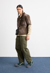 Camisa de cuero marrón de manga corta, capa larga beige, pantalones cargo verdes, zapatillas gris y verde; incluye un bolso cruzado negro.