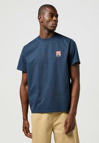 Marinblå bomullströja med korta ärmar och rund halsringning; har en liten fyrkantig grafisk logotyp på vänster bröstområde.