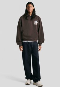 Felpa cappuccio marrone con tasca frontale e testo grafico bianco, abbinata a jeans a gamba larga blu scuro e scarpe da ginnastica chiare.