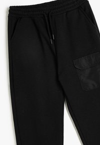 Schwarze Jogginghose mit elastischem Bund und Kordelzug, zwei Seitentaschen und einer strukturierten Stofftasche am Oberschenkel.