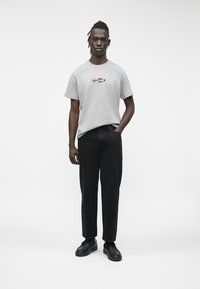 Šedé bavlněné tričko s červeným a černým logem "Guess Jeans", doplněné černými kalhotami a černými teniskami, stojící na jednobarevném bílém pozadí.