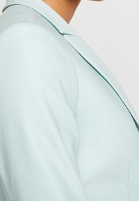 Ljusblå blazer med spetsig krage, slät textur och strukturerad passform, som visar en skräddarsydd design och rena linjer.