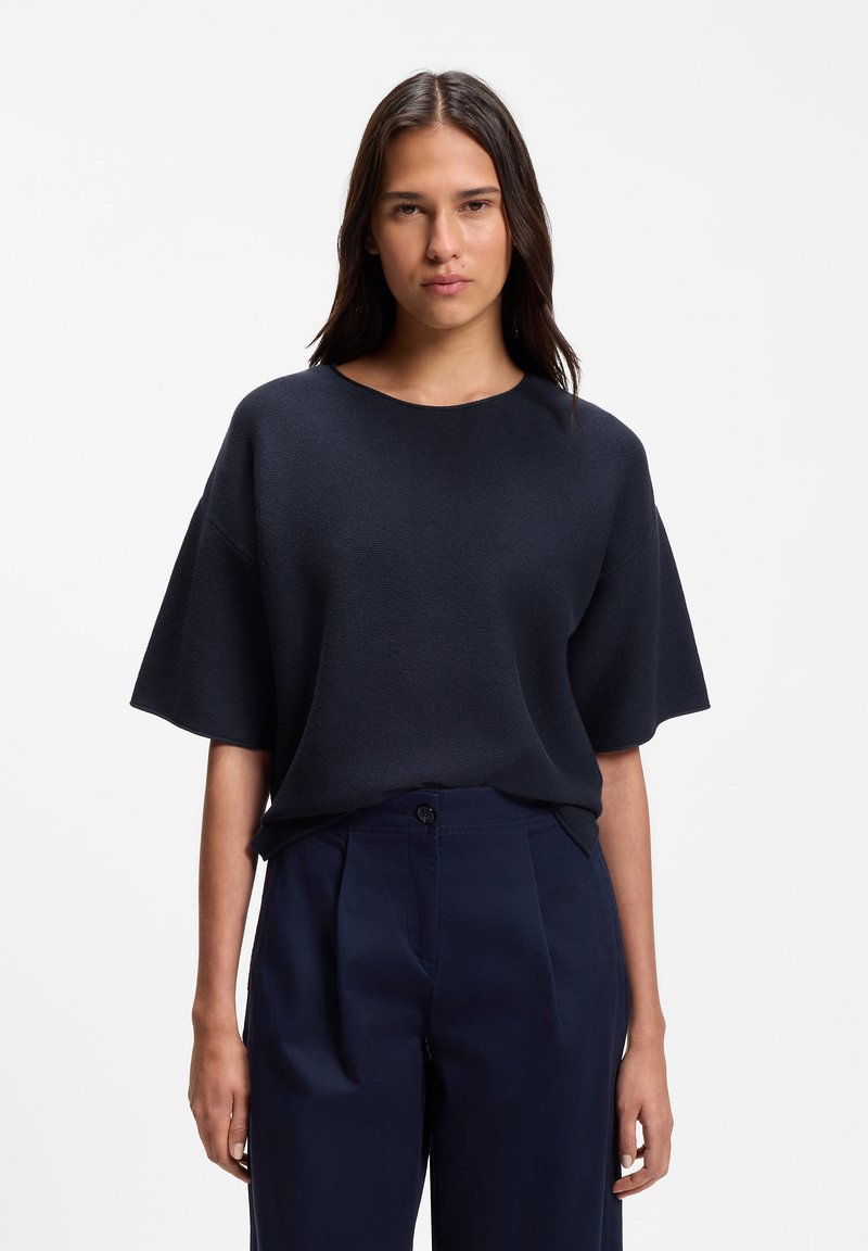 Femme aux cheveux raides et foncés portant un haut ample bleu marine foncé à manches courtes et un pantalon taille haute bleu marine sur fond blanc.