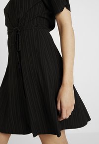 Schwarzes Kleid mit vertikalen Streifen, kurzen Ärmeln und einer gebundenen Taille sowie einem ausladenden Rock. Glatte Textur mit lässigen Designelementen.