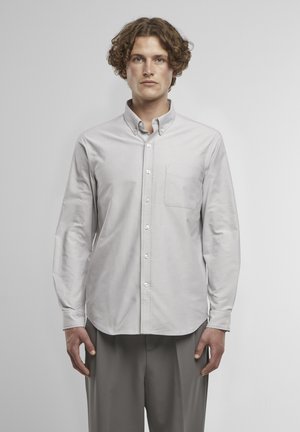 Junger Mann mit lockigem Haar, der ein hellgraues Hemd mit Knöpfen und Brusttasche sowie graue Hose trägt, vor einem schlichten Hintergrund steht.
