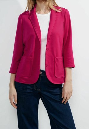 Femme portant un blazer rose vif avec deux poches avant, un haut blanc et un jean bleu foncé, debout devant un fond blanc uni.