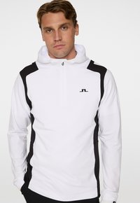 Vit kvartdragningströja med svarta sidopaneler och en logotyp på bröstet. Slät textur och justerbar huva. Enkel och funktionell design.