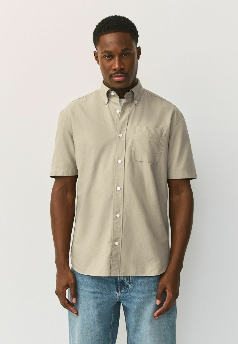 Homme debout devant un fond blanc, portant une chemise beige à manches courtes boutonnée et un jean bleu clair, regardant droit devant.
