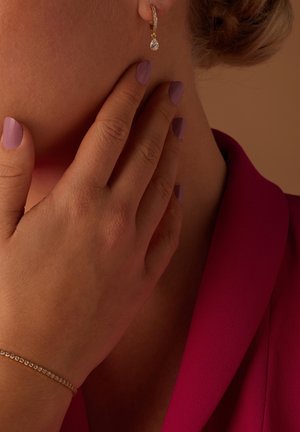 Boucle d'oreille en or avec un cristal en forme de goutte, assortie à une main présentant un vernis à ongles violet clair. L'arrière-plan est d'une teinte douce et neutre.