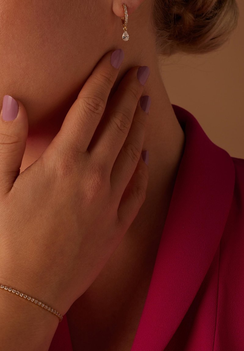 Gouden ringoorbel met een druppelvormige kristal, gecombineerd met een hand die lichtpaarse nagellak toont. De achtergrond heeft een zachte, neutrale tint.