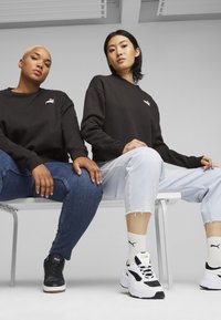 Felpe nere con loghi bianchi, jeans skinny blu e pantaloni cropped in denim chiaro, abbinati a sneakers nere e bianche, seduti su una panchina.