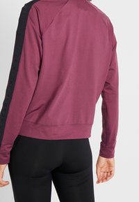 Bordeaux-rotes Sport-Sweatshirt mit schwarzem Logo-Detail an den Ärmeln, taillierte Passform, gerippter Saum und glatte Textur, kombiniert mit schwarzen Leggings.