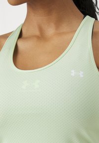Gros plan d'une personne portant un débardeur de sport perforé vert clair avec une sangle noire en dessous et un logo Under Armour blanc.