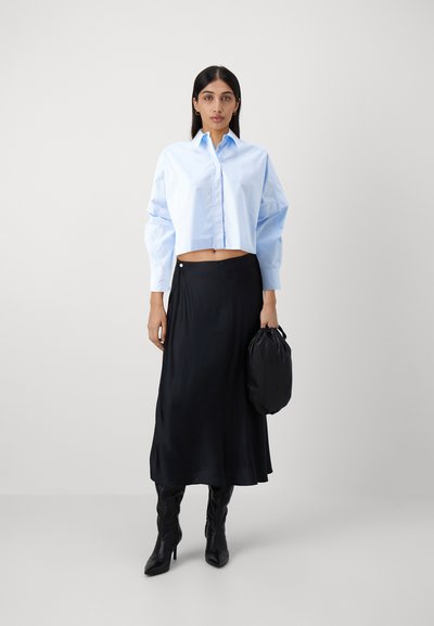 Camicia azzurra chiara cropped con maniche oversize, abbinata a una maxi gonna nera in satin e stivali neri con tacco, che tiene una borsa a pochette nera.