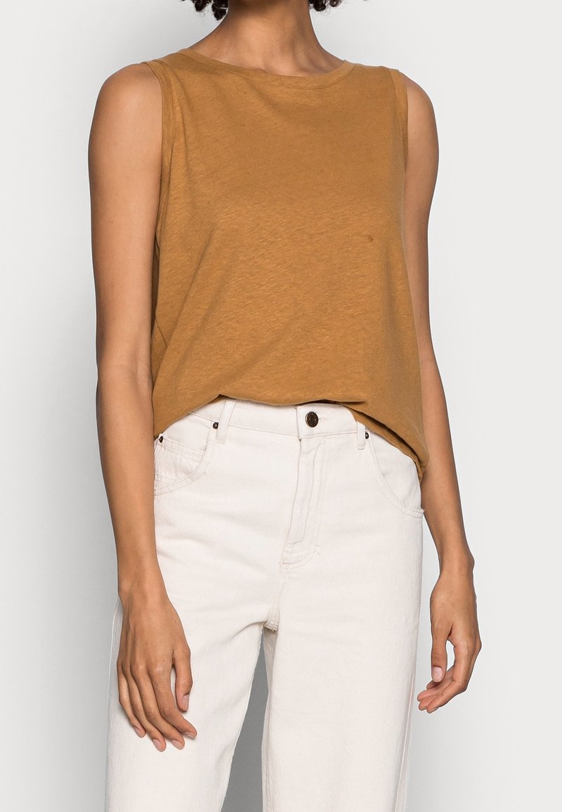 Mosterdgele mouwloze top van zacht katoen, gecombineerd met hoge witte jeans met knoopsluiting en zakken.