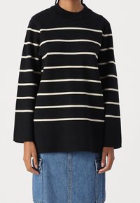 Personne portant un pull noir à manches longues avec des rayures horizontales blanches et une jupe en jean bleue avec de grandes poches.