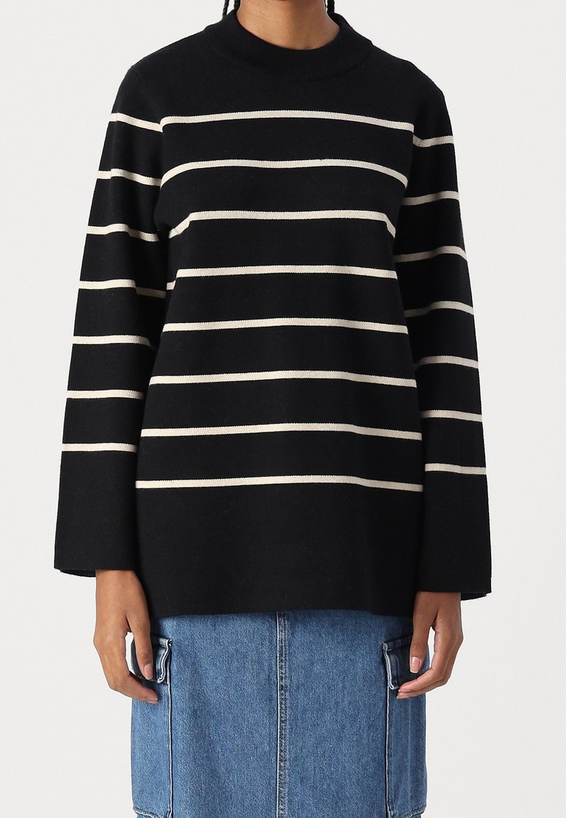 Personne portant un pull noir à manches longues avec des rayures horizontales blanches et une jupe en jean bleue avec de grandes poches.