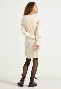 Kremowa sukienka z przezroczystymi rękawami, ozdobiona wzorem w białe kropki i dopasowanym krojem. Połączona z burgundowymi botkami na sznurówki.