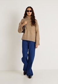 Bēša platā augstā kakla džemperis ar ribveida aprocēm, kombinēts ar plašām zilām džinsa biksēm, melnām saulesbrillēm un mazu melnu somiņu.