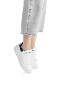 Zapatillas blancas con parte superior de piel suave y acento negro en el talón. Presenta puntera redonda, cordones planos y una suela blanca gruesa.