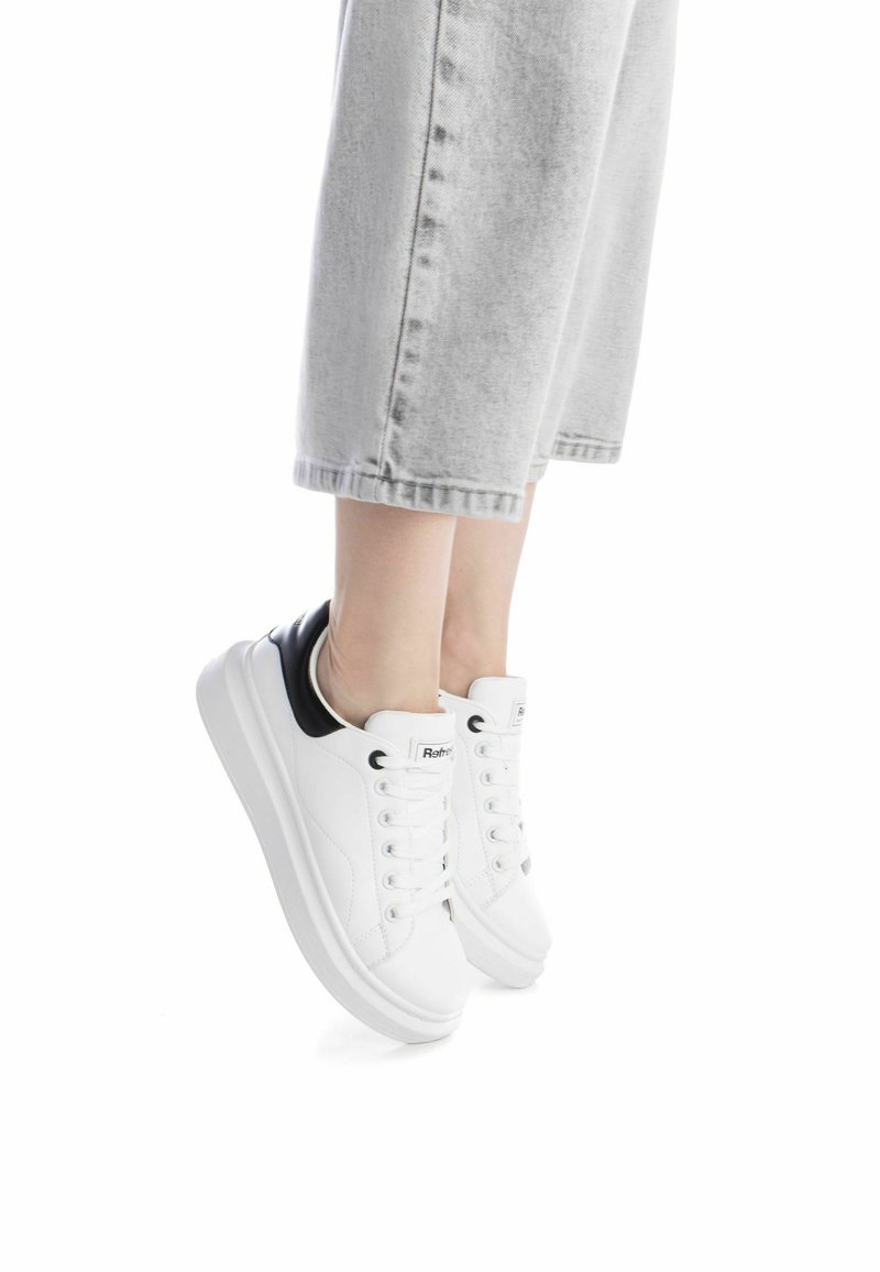 Zapatillas blancas con parte superior de piel suave y acento negro en el talón. Presenta puntera redonda, cordones planos y una suela blanca gruesa.