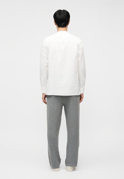 Άνδρας με την πλάτη γυρισμένη φοράει λευκό πουκάμισο με μακριά μανίκια, γκρι φαρδιά παντελόνια και λευκά αθλητικά παπούτσια, σε απλό ανοιχτό φόντο.