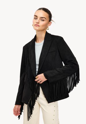 Mujer con el cabello recogido en un moño que lleva una chaqueta de ante negra con flecos sobre una blusa gris claro y pantalones crema con tachuelas doradas.