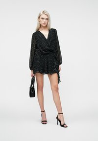 Melnā aptīta kleita ar caurspīdīgām garām piedurknēm, punktiņu raksts, viļņota apakšdaļa, valkāta ar strappy papēžiem un mazu melnu rokassomu.