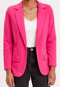 Blazer rose fuchsia avec un col cranté, deux poches avant et des manches longues. Porté sur un haut blanc orné de dentelle et un jean noir.