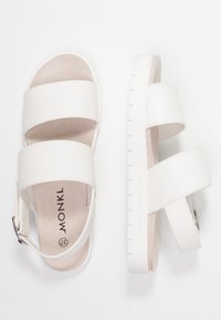 Bílé sandály s dvěma širokými texturovanými popruhy, polstrovanou stélkou a gumovou podrážkou s mírným vzorem pro lepší přilnavost. Nastavitelná spona po straně.