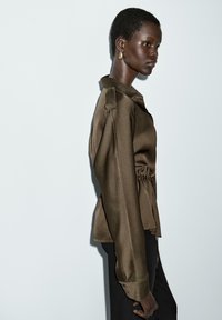 Blouse en satin marron avec un col, des manches longues et un détail froncé sur le côté, associée à un pantalon noir, présentée de profil sur un fond clair.
