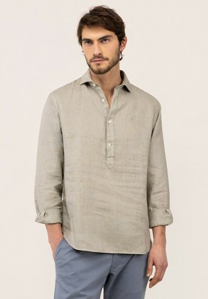 Hombre con camisa de lino beige de manga larga con puños arremangados y pantalones azules, de pie frente a un fondo liso y claro.