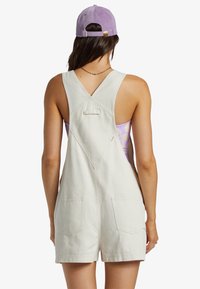 Salopette in denim beige chiaro con design a V sul retro, dotata di due tasche posteriori e spalline regolabili, indossata sopra una maglietta viola.