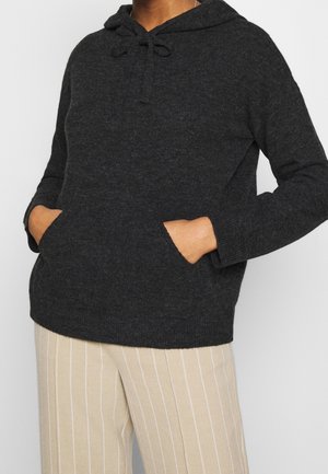 Personne portant un sweat à capuche en tricot gris foncé avec poches avant et un pantalon beige à fines rayures blanches verticales, mains dans les poches.