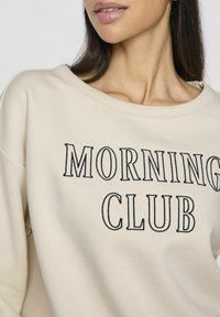 Sudadera color crema con un escote amplio, que presenta "MORNING CLUB" bordado en azul oscuro. Tejido suave con un ajuste relajado.