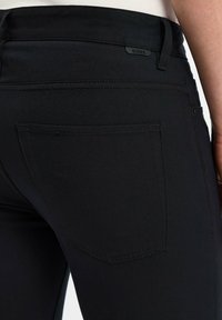 Zbliżenie na kieszeń czarnych dopasowanych spodni z widocznym szyciem i małą metką "BOSS" blisko pasa na biodrze osoby.