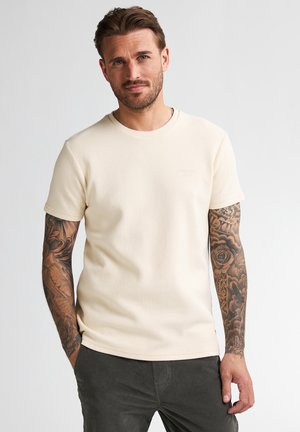 Mann mit kurzem braunem Haar, der ein cremefarbenes kurzärmeliges Hemd und dunkle Hosen trägt, zeigt detaillierte Tattoos auf beiden Armen.