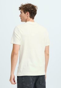 Light beige short-sleeve t-shirt with a round neck, made from soft cotton. The back features minimal design and a small logo tag near the hem.
