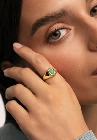 Anillo de oro con una piedra preciosa verde ovalada, rodeada de pequeños acentos negros, llevado en una mano con manicura rosa claro.