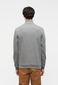 Γκρι φούτερ με ψηλό γιακά, μακριά μανίκια και ριμπ μανσέτες, παρουσιασμένο σε μοντέλο που κοιτάζει προς τα πίσω μπροστά σε ουδέτερο φόντο.