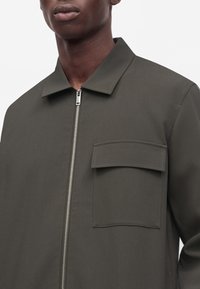Homme portant une veste vert olive foncé avec une fermeture éclair métallique à l'avant et une grande poche à rabat sur la poitrine gauche, visible du cou jusqu'au milieu du torse.