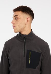 Grijze half-zip pullover van gestructureerde stof, met een zwarte borstzak en een gele logoaccent. Eenvoudig, functioneel ontwerp.