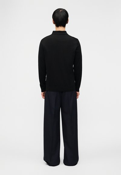 Άτομο που στέκεται με την πλάτη προς την κάμερα, φορώντας μαύρο μπλουζάκι μακρυμάνικο και φαρδιά σκούρα παντελόνια μπροστά σε ένα απλό φωτεινό φόντο.