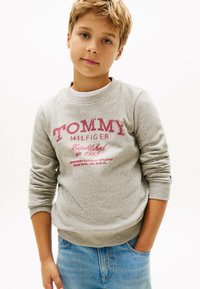 Szara bluza z logo "TOMMY HILFIGER" w kolorze czerwonym, z prążkowanymi mankietami i dołem. Nosi się ją na białej koszuli, w zestawieniu z jasnoniebieskimi dżinsami.