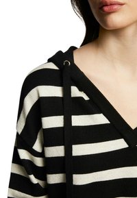 Femme portant un sweat à capuche rayé noir et blanc avec des cordons, montrant une partie de son visage et de son cou sur fond blanc.