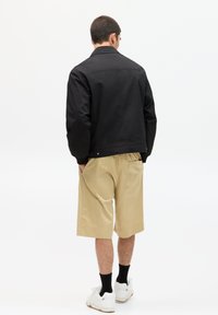 Schwarze Jacke mit Kragen, Rückennahtdetail, kombiniert mit beigen Shorts. Das Outfit wird mit weißen Sneakers und schwarzen Socken ergänzt.
