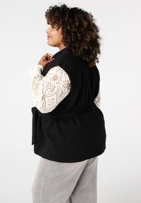 Zwarte gewatteerde vest met een strikriem, gedragen over een gebloemde ivoorkleurige blouse met lange mouwen. Zachte textuur en een relaxed fit. Lichtgrijze broek.